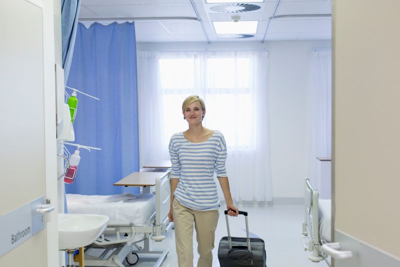 Eine Frau verlässt glücklich ein Krankenhauszimmer mit ihrem Koffer.