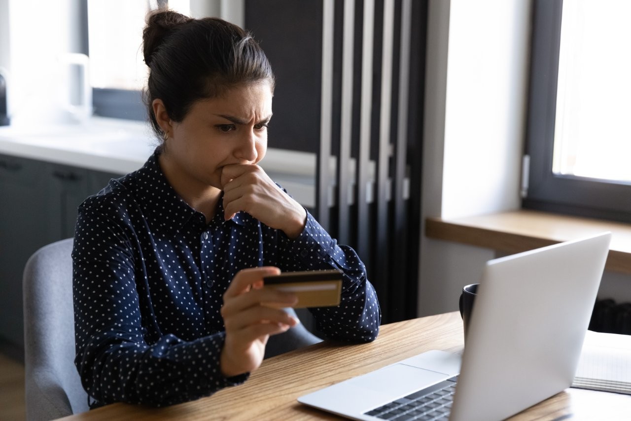 A woman sits in front of her laptop and looks sceptically at her credit card.