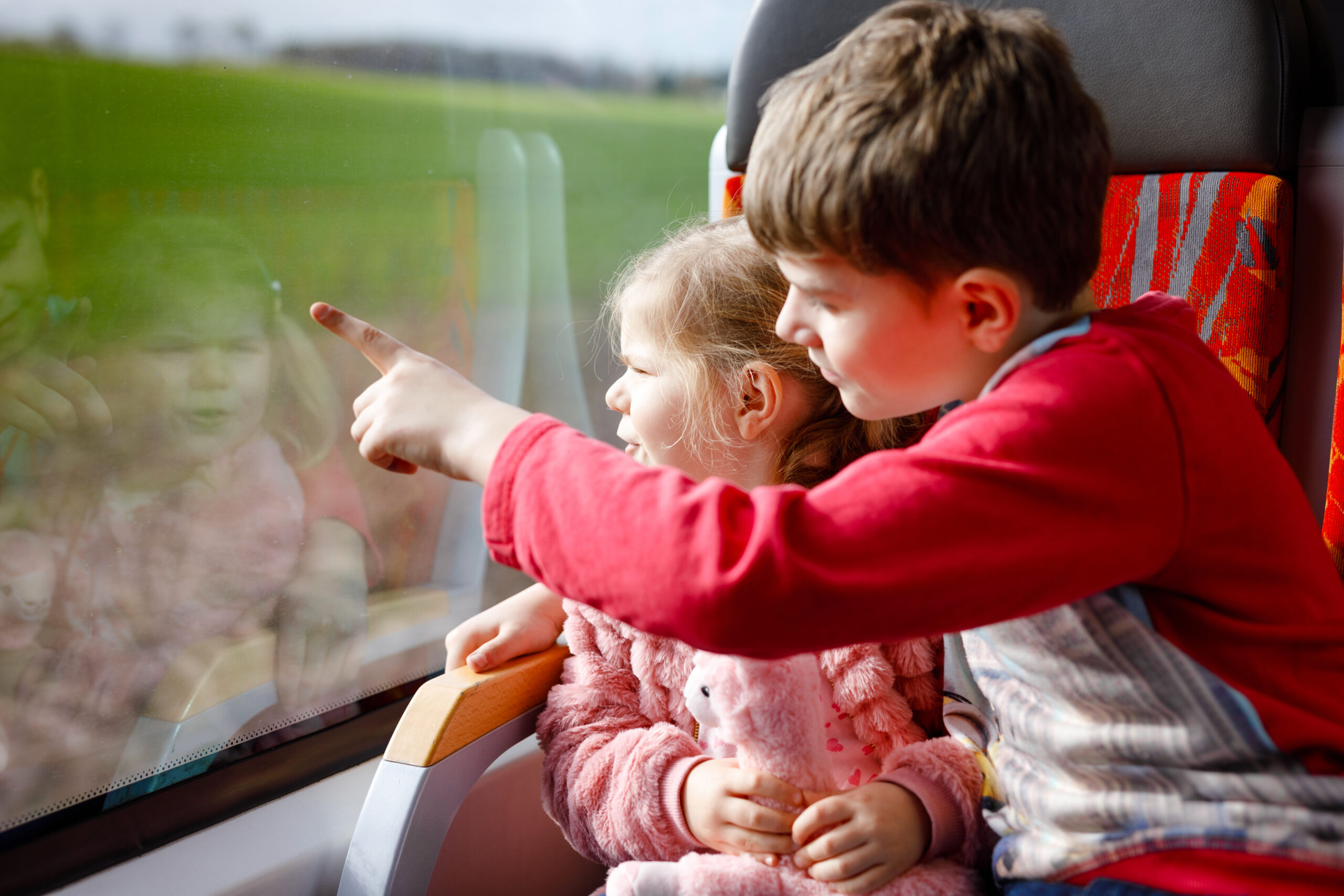Two children look out the window of a train while the boy shows the girl something outside