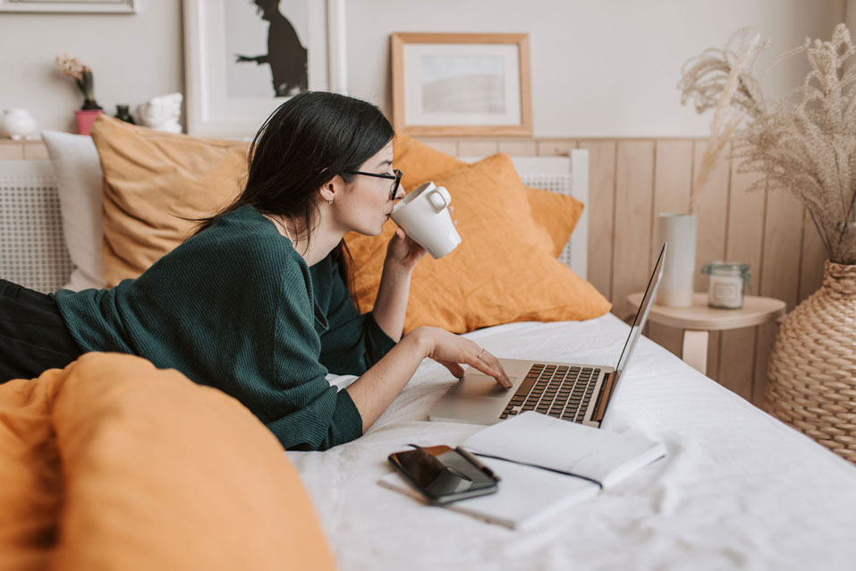 A woman is lying on the bed, working on her laptop and drinking from a cup. Next to her is a smartphone on a notebook.