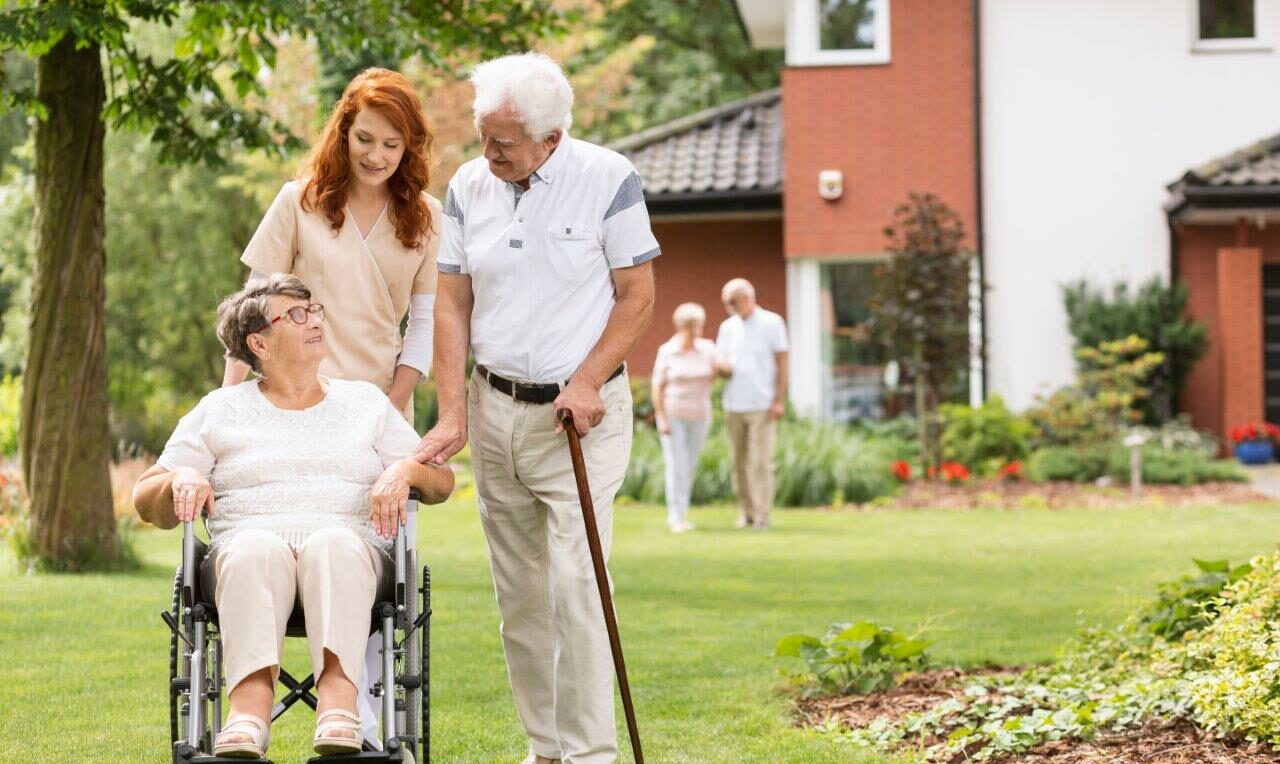 Eine ältere Dame im Rollstuhl beim Spaziergang im Garten. Sie wird begleitet von einer jungen Pflegerin mit roten Haaren und einem älteren Herren mit Gehstock.